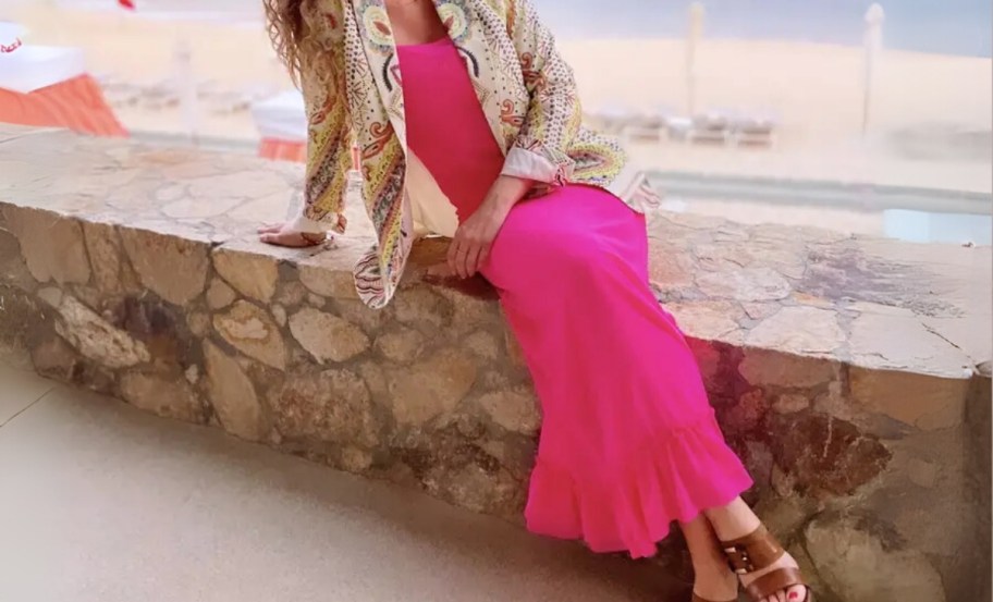 a woman sitting by the coastline in a hot pink maxidress and jacket