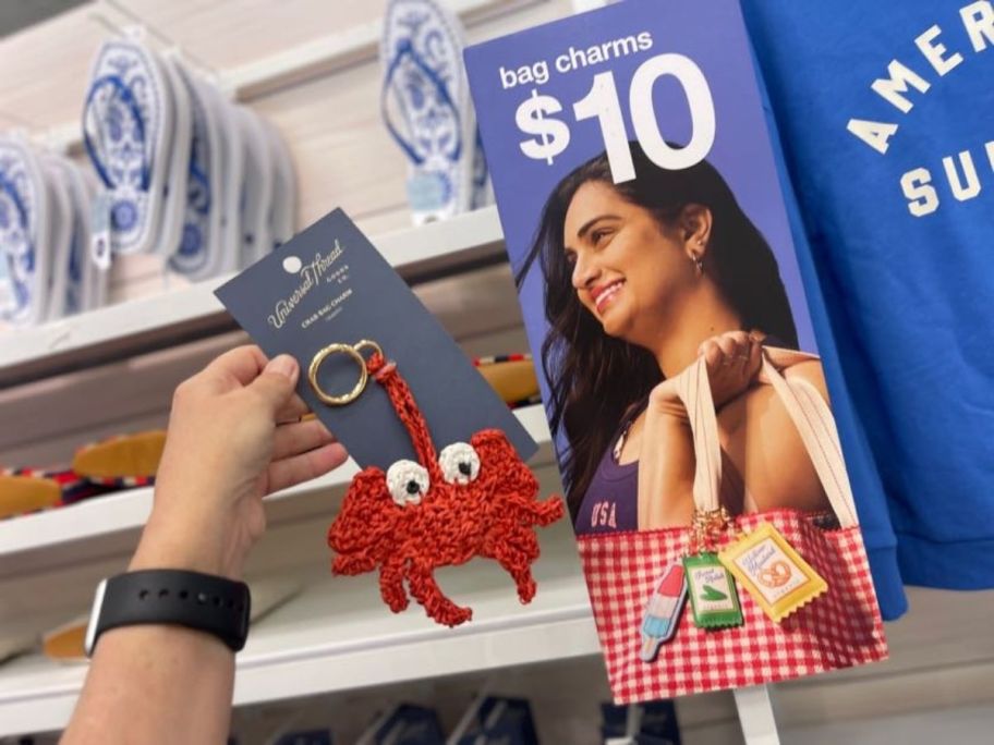 person holding up crab bag charm next to bag charms sign