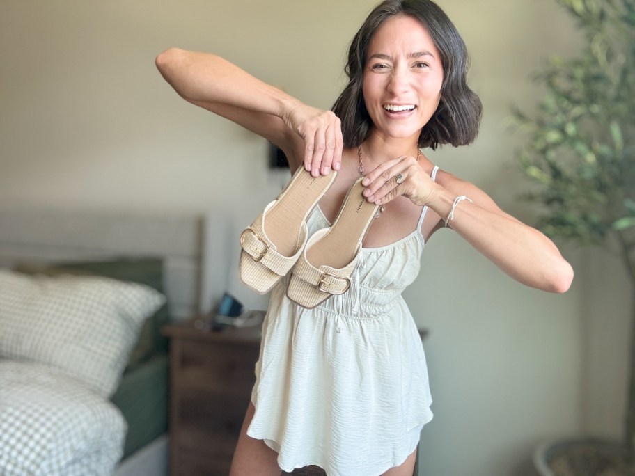 smiling woman holding a pair of shoes in front of her body with her elbows out at her sides