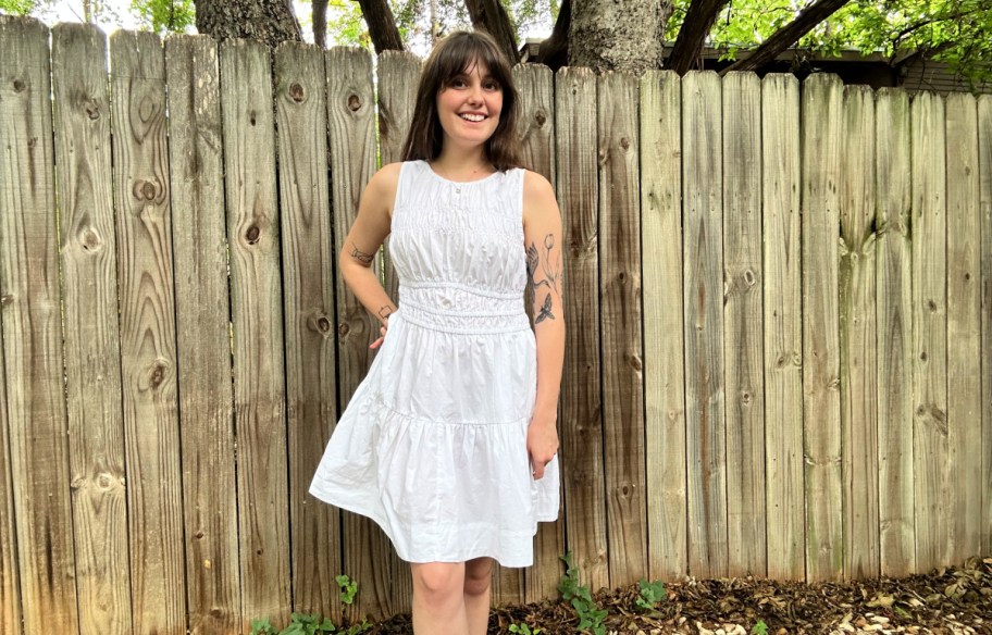 smiling woman in a white dress standing in front of a wood fence outside