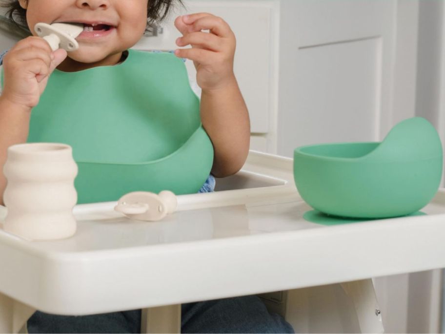 A baby in a high chair with a silicone bib, bowl, cup and spoons