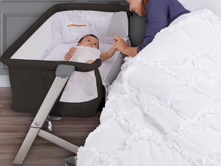 A mom touching a baby inside of a Dream On Me Skylar Bassinet & Beside Sleeper next to a bed