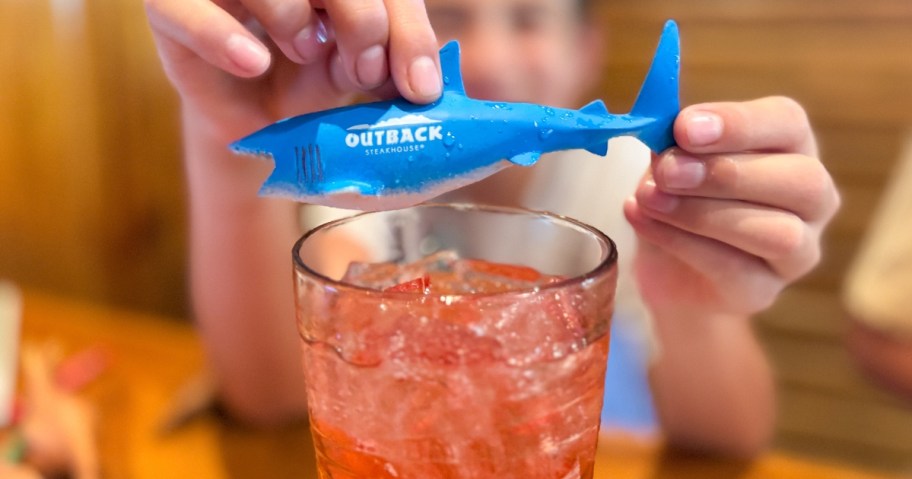 kid holding a plastic toy shark above a glass with a red drink in it