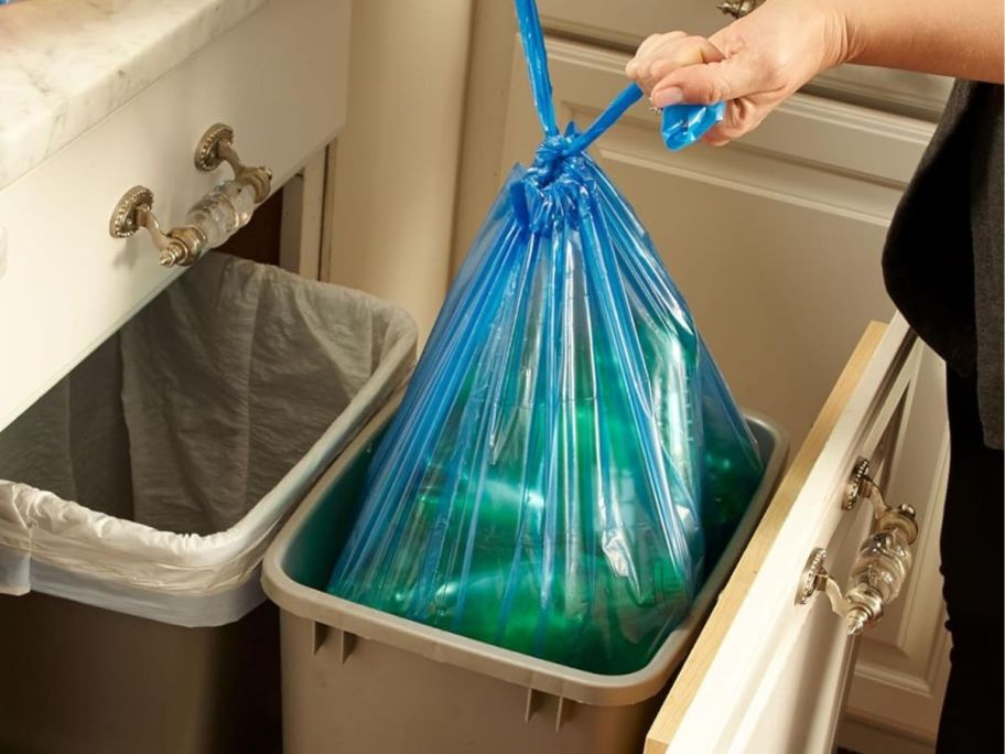 A person tying up a blue Hefty trash bag in a kitchen in a trash bin