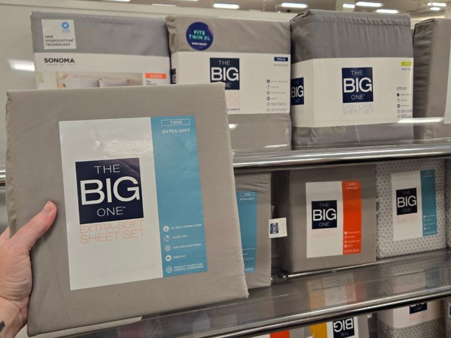 Hand holding up a pack of Kohl's The Big One Extra Soft Sheets next to shelves filled with more of them