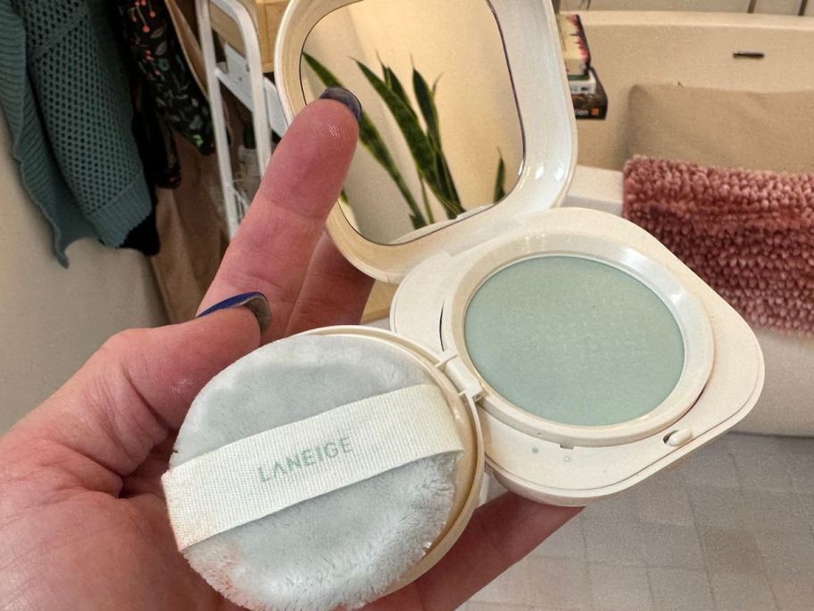 hand holding compact opened with puff and green blurrying powder