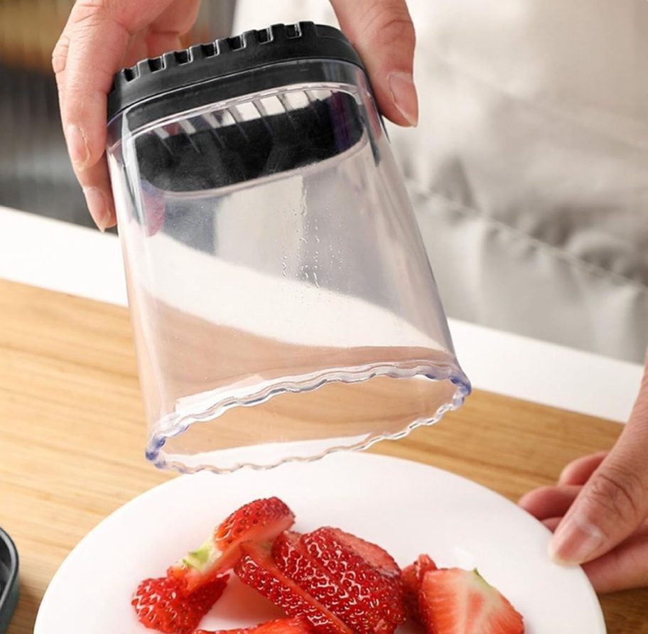 a womans hand holding a fruit slicer cup buy the bottom as she dumps sliced strawberries onto a plate