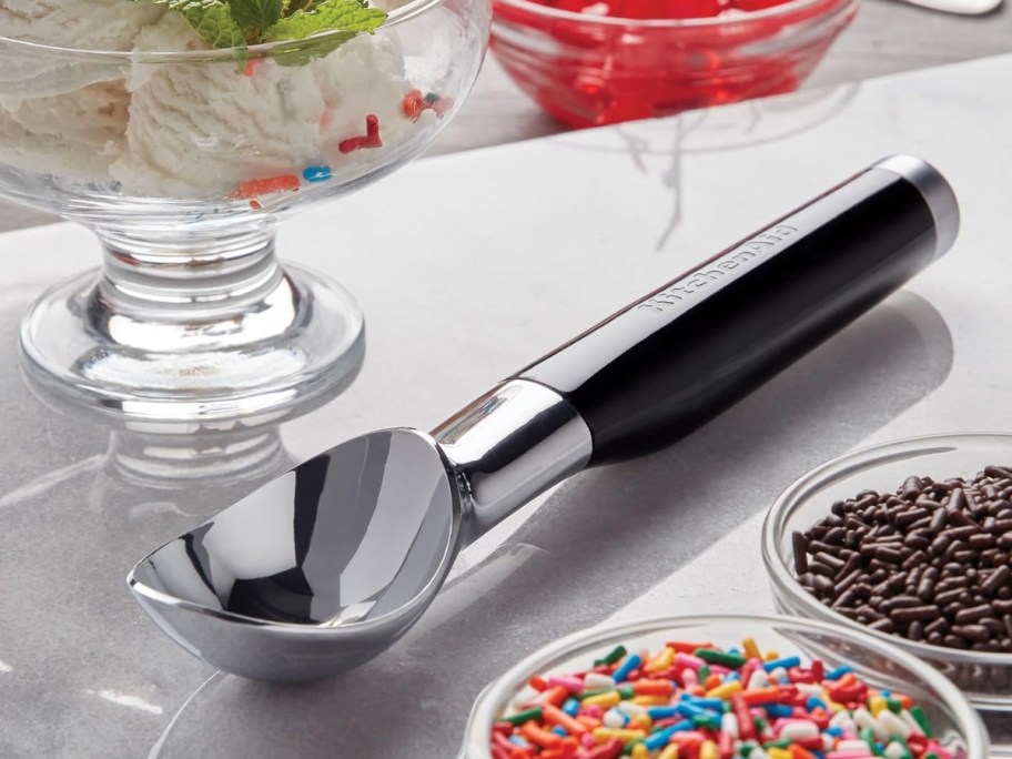 a KitchenAid ice cream scoop next to a bowl of ice cream and bowls of toppings
