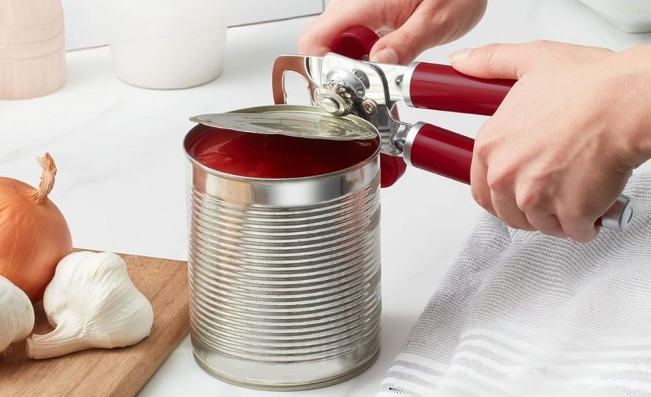 a womans hand using a red can opener to open a can of tomato sauce