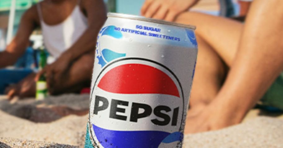 pepsi prebiotic soda can in sand on beach with people behind it